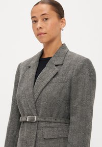 Blazer de padrão pied-de-poule em cinza e preto, com cintura cativada, lapelas recortadas e uma silhueta estruturada. Acabamento de tecido suave.