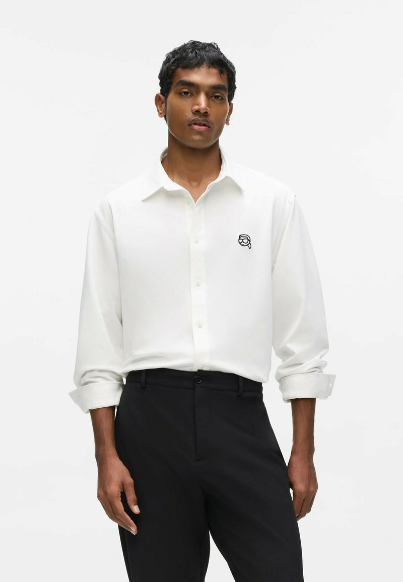 Homme portant une chemise blanche boutonnée avec un petit logo noir sur la poitrine et un pantalon noir, debout devant un fond blanc uni.