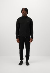 Schwarze Zip-Jacke mit hohem Kragen und Logo, kombiniert mit passenden schwarzen Jogginghosen. Beide Kleidungsstücke haben eine glatte Textur und eine taillierte Passform.