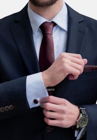 Mornarica sako s belim košuljom, kravatom s burgundskim dijagonalnim karirama i manžetnim dugmadi. Detalji uključuju srebrni sat i maramicu u džepu koja se poklapa s kravatom.