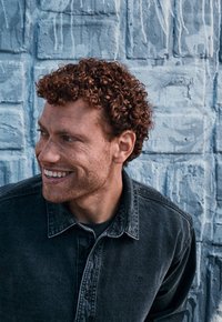 Zwarte denim knopenoverhemd op een persoon met krullend rood haar, glimlachend tegen een blauwe gestructureerde muur als achtergrond.
