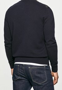 Navyfarbener gestrickter Pullover mit geripptem Saum und Bündchen, verziert mit strukturierten Schulterakzenten. Kombiniert mit dunkelblauen Jeans.