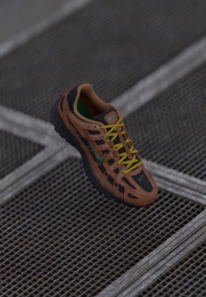 Brun og sort Nike sneaker med gule snørebånd, der hviler på gråt metalristegulv.