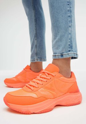 Person wearing bright orange chunky sneakers and light blue rolled-up jeans, standing on a white surface.