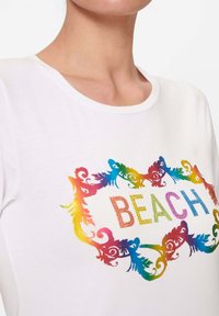 White short-sleeve t-shirt featuring a colorful, gradient "BEACH" text embellished with rhinestones in a decorative frame design.
