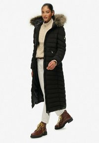 Cappotto impermeabile nero con cappuccio foderato in pelliccia, chiusura con zip, texture a righe verticali, abbinato a un maglione beige e pantaloni crema, con stivali marroni.