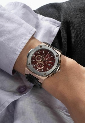 Silver and rose gold watch with a red dial, chronograph features, and black leather strap, worn on a wrist under a light purple shirt.
