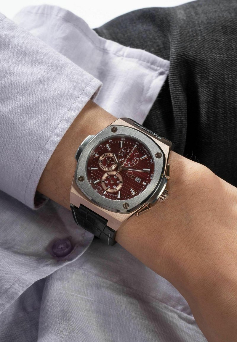 Orologio in argento e oro rosa con un quadrante rosso, funzioni cronografo e cinturino in pelle nera, indossato su un polso sotto una camicia di colore lilla.