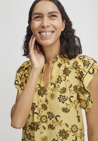 Blusa floreale gialla con maniche corte, caratterizzata da un neckline a chiave, motivi floreali neri e un tessuto morbido e texturizzato.