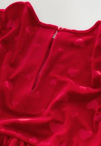 Vestito rosso con una morbida texture vellutata e un motivo a cuori su tutta la superficie. Presenta un'apertura a chiave sul retro, fissata con un bottone rosa.