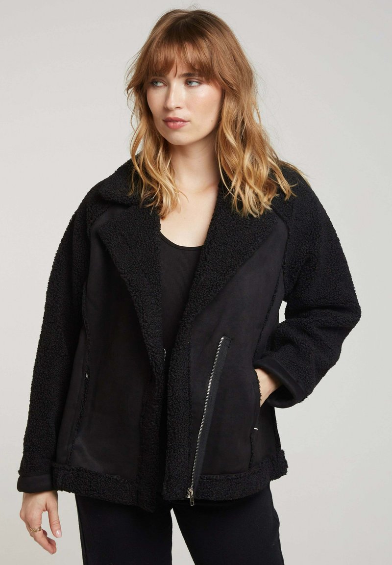 Veste d'extérieur noire avec une texture pelucheuse, un col surdimensionné et deux poches frontales avec fermeture éclair. Tissu doux avec des finitions contrastées.