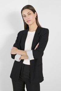 Melna piegriezta žakete ar sarullētām svītrām, viegla auduma, divas priekšējās kabatas, kopā ar vienkāršu baltu blūzi un melnām biksēm.
