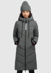 Manteau long matelassé gris avec fermeture zippée, capuche et motif en chevrons. Présente des poches latérales avec des accents noirs. Porté avec un bonnet en tricot assorti.