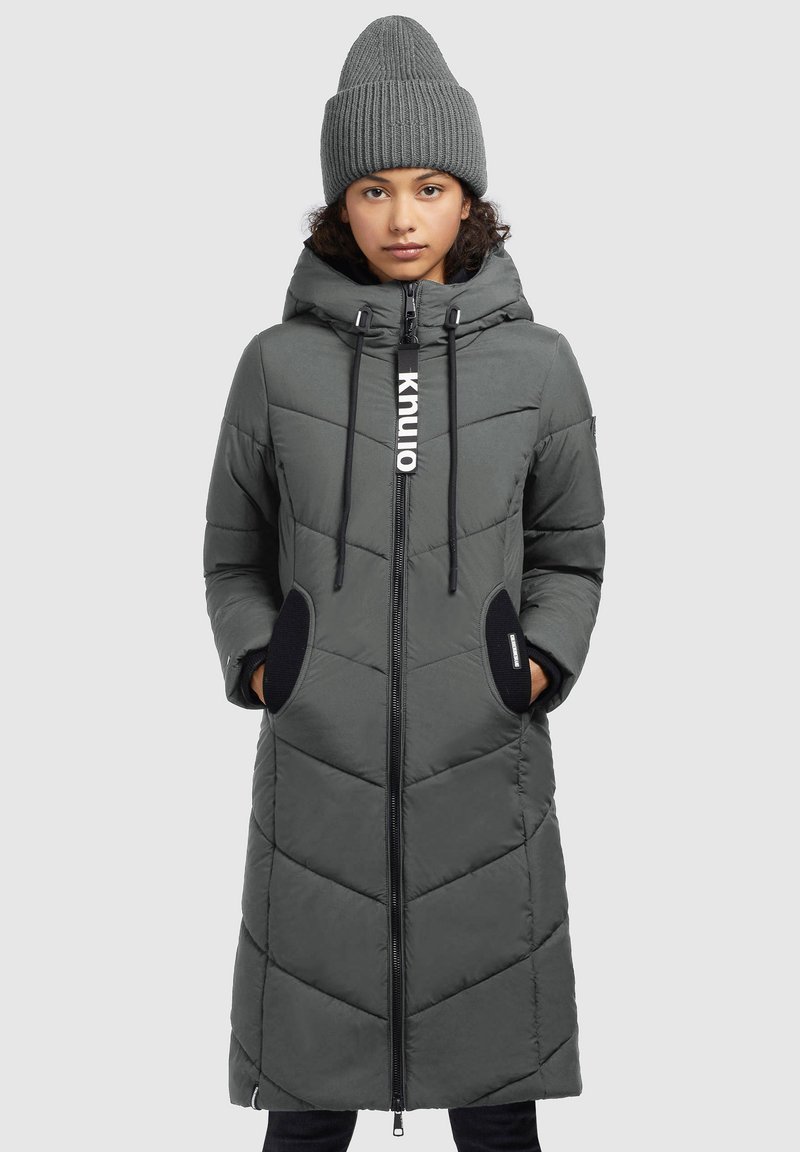 Manteau long matelassé gris avec fermeture zippée, capuche et motif en chevrons. Présente des poches latérales avec des accents noirs. Porté avec un bonnet en tricot assorti.