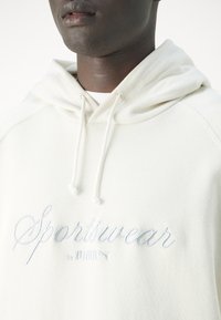 Světle béžová mikina s klokaní kapsou, kapucí se stahovacími šňůrkami a vyšitým textem "Sportswear by PUMA" ve světle modré barvě. Měkká textura.