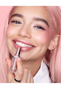 Lip gloss in a clear tube with a silver applicator. Model has pink hair and a smile, showcasing glossy, light pink lips and flawless skin.