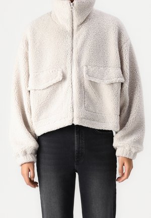 Kobieta ubrana w jasnobeżową, teksturowaną kurtkę polarową z przednimi kieszeniami na klapę i zamkiem błyskawicznym, zestawioną z czarnymi, wysokim stanem jeansami.