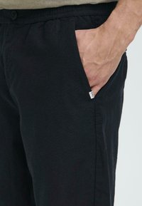 Svart bomullsbukser med elastisk midjebånd og en sidelomme. Har en liten merking nær lommeåpningen. Glatt tekstur.