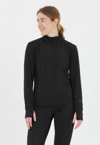 Sort atletisk jakke med høj krave, fuld lynlås, tekstureret stof og tommelhuller ved manchetterne. Bæres sammen med matchende sorte leggings.