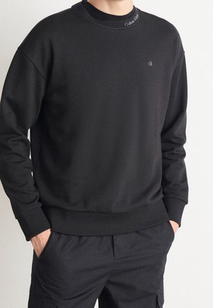 Mand iført en sort Calvin Klein sweatshirt med lille "ck" logo på brystet og "Calvin Klein" tekst på kraven, hænderne i bukselommerne.