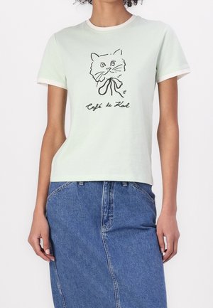 Kvinde iført en hvid T-shirt med en sort skitse af en kats ansigt og teksten "Café de Karl", kombineret med et blåt denimskørt.