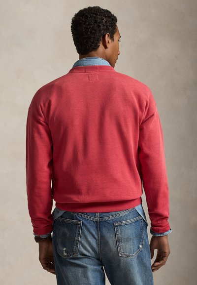 Άνδρας με την πλάτη στραμμένη, φοράει κοραλλί πουλόβερ με μακριά μανίκια πάνω από τζιν πουκάμισο και ξεθωριασμένο μπλε τζιν, με ουδέτερο φόντο.