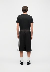 Άνδρας στέκεται με την πλάτη γυρισμένη, φοράει μαύρο μπλουζάκι, μαύρα σορτς μέχρι το γόνατο με λευκές λωρίδες στο πλάι, λευκές κάλτσες και μαύρα αθλητικά παπούτσια σε λευκό φόντο.