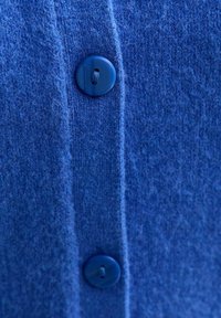 Close-up van een blauwe wollen cardigan, met een gestructureerde stof, verticale ribbels en twee bijpassende blauwe knopen langs de split.