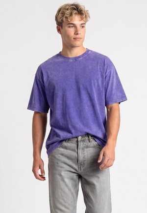 Giovane uomo con capelli biondi ricci che indossa una maglietta viola larga e jeans grigio chiaro, in piedi contro uno sfondo semplice.
