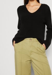 Frau trägt einen schwarzen V-Ausschnitt-Strickpullover, der in hochgeschnittene beigetöne Hosen gesteckt ist, mit Händen in den Taschen vor einem schlichten Hintergrund.