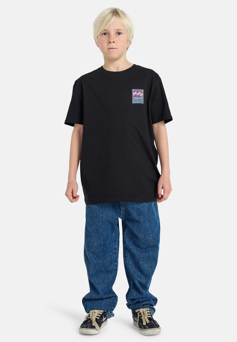 Black cotton t-shirt with short sleeves and a small colorful logo patch on the left chest, paired with loose blue denim jeans and black shoes.