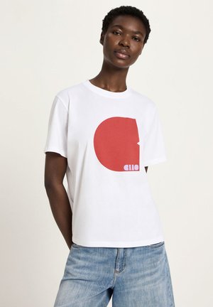 Woman with short curly hair wearing a white t-shirt with a large red abstract shape and "ciao" text, paired with light blue jeans.