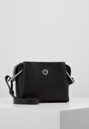 Sort tekstureret læder crossbody taske med justerbar rem og sølv rundt logoemblem placeret i midten af frontpanelet.