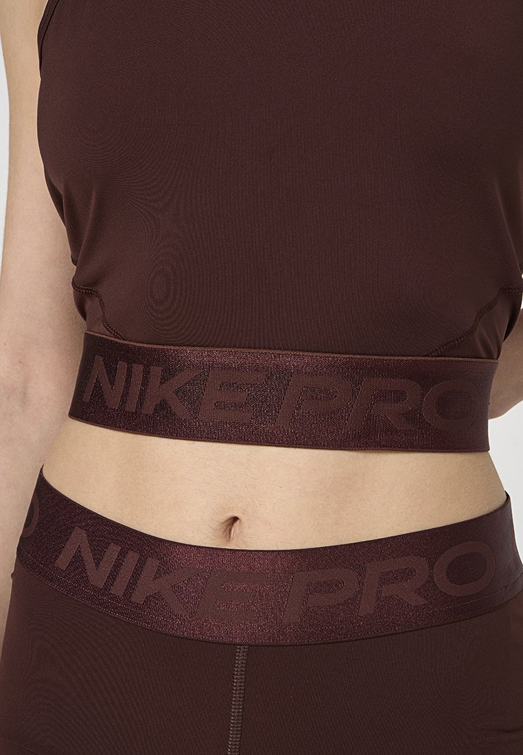 Haut de sport Nike Pro bordeaux avec une bande brillante arborant le nom de la marque. Tissu lisse et extensible avec des coutures plates pour le confort et l'ajustement.