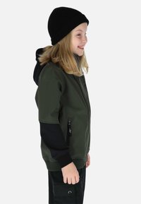 Smilende barn iført en sort hue, mørkegrøn jakke med sorte ærmer og sorte cargobukser, vendt mod højre på en lys baggrund.