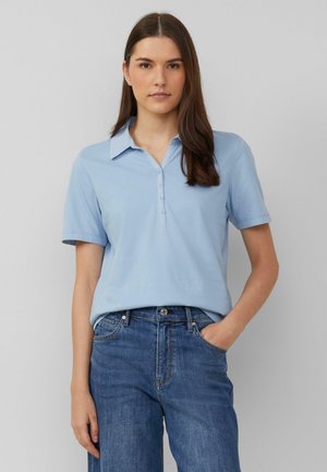 Lichtblauwe polo met een kraag, met korte mouwen en een knoopsluiting. Draag met denim jeans, handen in de zakken.