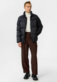 Doudoune noire avec un col montant, portée sur un col roulé crème. Un pantalon plissé marron et des chaussures noires à lacets complètent la tenue.