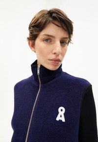 Marine blaue Fleeceweste mit hohem Kragen, silbernem Reißverschluss, schwarzen Akzenten und weißem gesticktem Logo auf der linken Brust. Glatte Textur, stylisches Design.