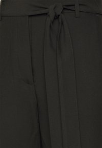 Μαύρο παντελόνι από ύφασμα με ζώνη που δένει στη μέση, με επίπεδο μπροστινό μέρος, πλαϊνές τσέπες και λεία υφή. Ο σχεδιασμός είναι απλός και λειτουργικός.