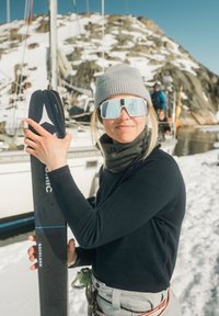 Ski with a dark top sheet and blue accents held by a person in a gray beanie and sunglasses, with snow and boats in the background.