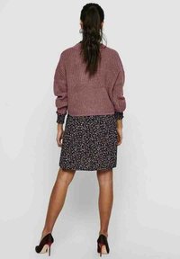 Gehaakte trui in zacht mauve, gecombineerd met een zwarte bloemenjurk. De outfit heeft lange mouwen en hoge hakken met een rode zool.