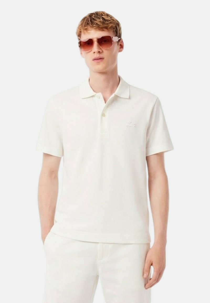 Jonge man die een wit poloshirt en een broek draagt met vierkante zonnebrillen met roze tint, tegen een effen achtergrond.