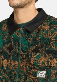 Groen en bruin fleecejack met ritssluiting en een corduroy kraag, gemaakt van een gestructureerde stof en met een logopatch aan de voorkant.