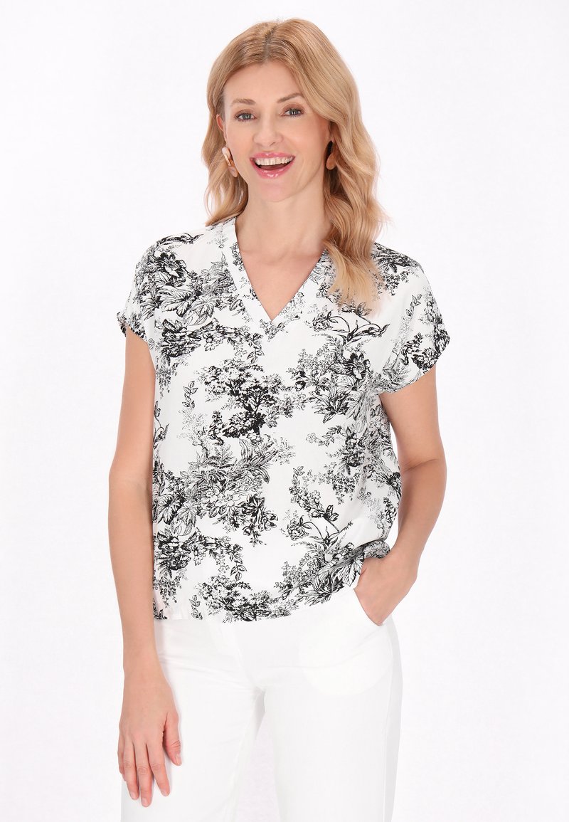 Blusa a maniche corte in tessuto bianco con motivo floreale nero. Presenta una scollatura a V e una vestibilità rilassata. Indossata con pantaloni bianchi.