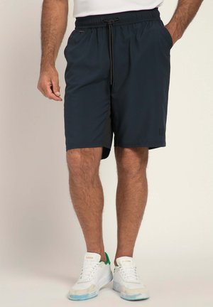 Mężczyzna w czarnych szortach na sznurek i białych butach Adidas z zielonymi akcentami, stojący z jedną ręką w kieszeni.