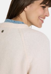 Hellrosa Pullover mit weicher Textur, runder Halslinie und einem kleinen metallischen Akzent auf der Rückseite. Sichtbarer blauer Denimkragen.