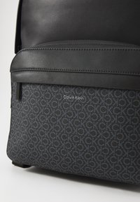 Zaino nero con dettagli in pelle liscia e un tessuto texture con un motivo "CK" ripetuto. Include una tasca frontale con zip.