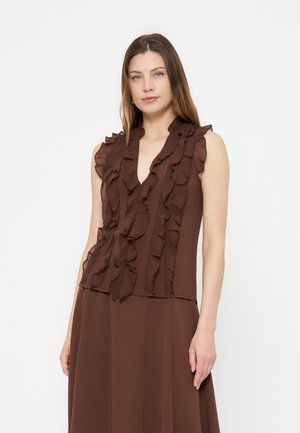 Femme portant un chemisier marron sans manches à volants superposés et une jupe marron assortie, debout sur un fond blanc uni.