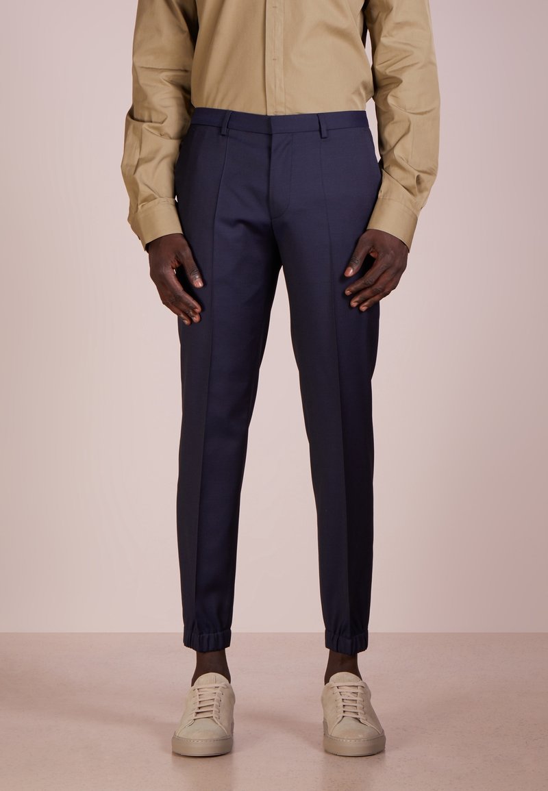 Marina tapered byxor med elastiska muddar, slätt tyg, sydda sömmar, bärs med en beige långärmad skjorta och ljusa sneakers.