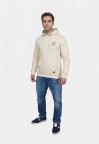 Kremowa bluza z kapturem z przednią kieszenią, z okrągłym logo na klatce piersiowej. Połączona z niebieskimi dżinsami i granatowymi trampkami.